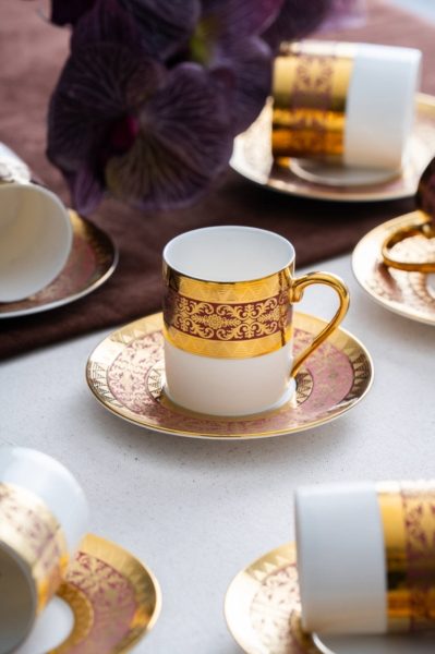 Kulîlkek spî bi xemilandina zêr û borgundî ya fireh li ser tasek lihevhatî ya ku bi qedehên wekhev dorpêçkirî ye radiweste.