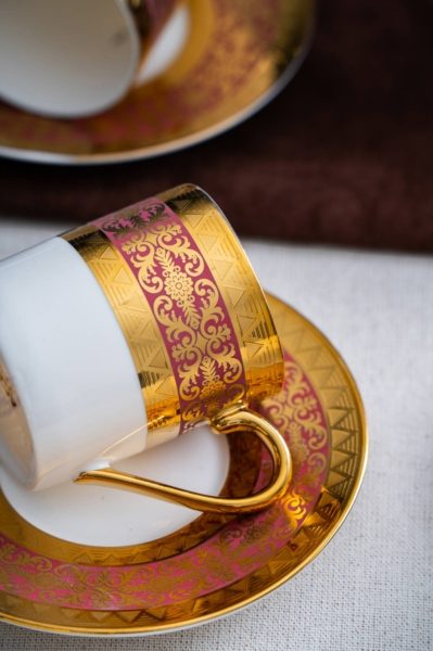 Kulîlkek porselenê ya spî ya bi qalibekî bordûnî û zêr li kêleka xwe li ser tasek lihevhatî radiweste.