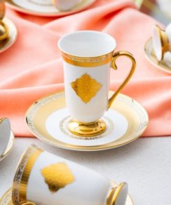 Elegant kaffekopp med gullkant og dekor på fat, plassert på en rosa duk.