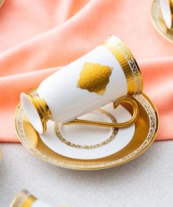 En elegant hvit og gullfarget kopp med skål ligger på siden på et bord dekket med et ferskenfarget klede.