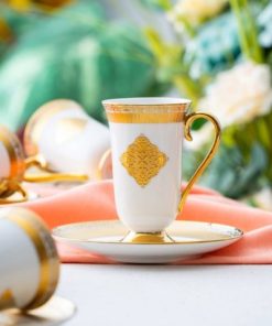 En elegant hvit kopp med gullmønster står på en matchende skål foran et ferskenfarget klede og blomster i bakgrunnen.