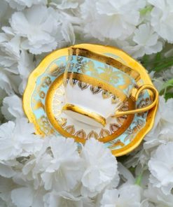 En elegant hvit og gullfarget kopp med blå detaljer ligger på en matchende skål omgitt av hvite blomster.