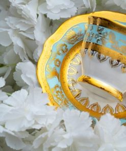 En elegant hvit porselenskopp med gull- og blåmønster ligger på en matchende skål omgitt av hvite blomster.