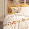 Oppredd seng med hvitt sengetøy dekorert med broderte blomster i gule, røde og blå farger. Putene og dynen har matchende blomstermønstre. Bakgrunnen består av en steinvegg med en terrakottakrukke med røde blomster, samt en trepersienne og beige veggpanel."