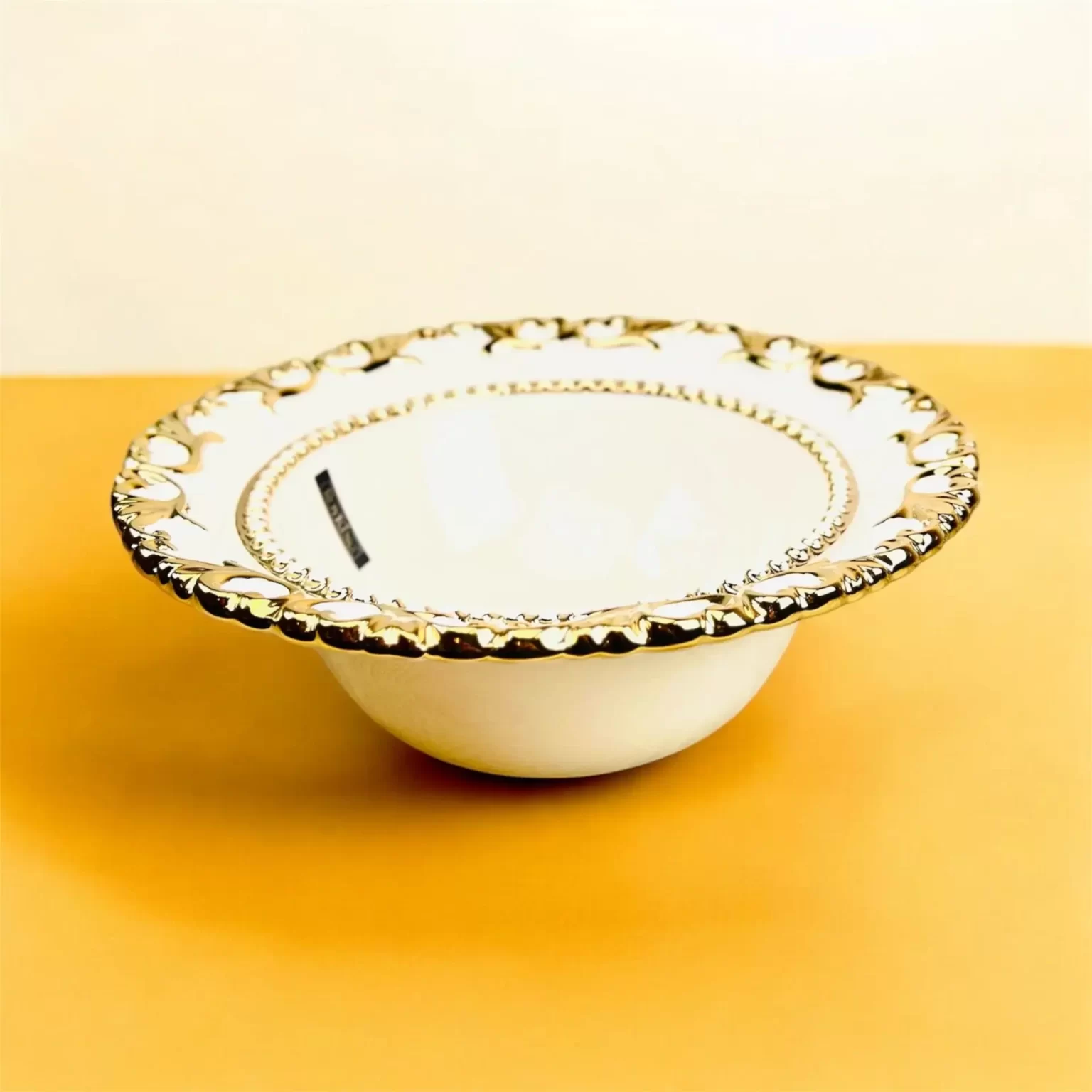 Elegant, rund serveringsskål i hvid keramik med en dekorativ guldkant, placeret på orange baggrund.