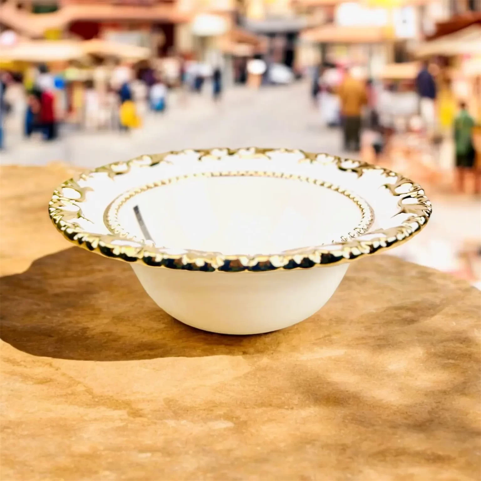 Dekorativ hvid serveringsskål med guldkant placeret på stenoverfladen, med en sløret markedsscene i baggrunden.