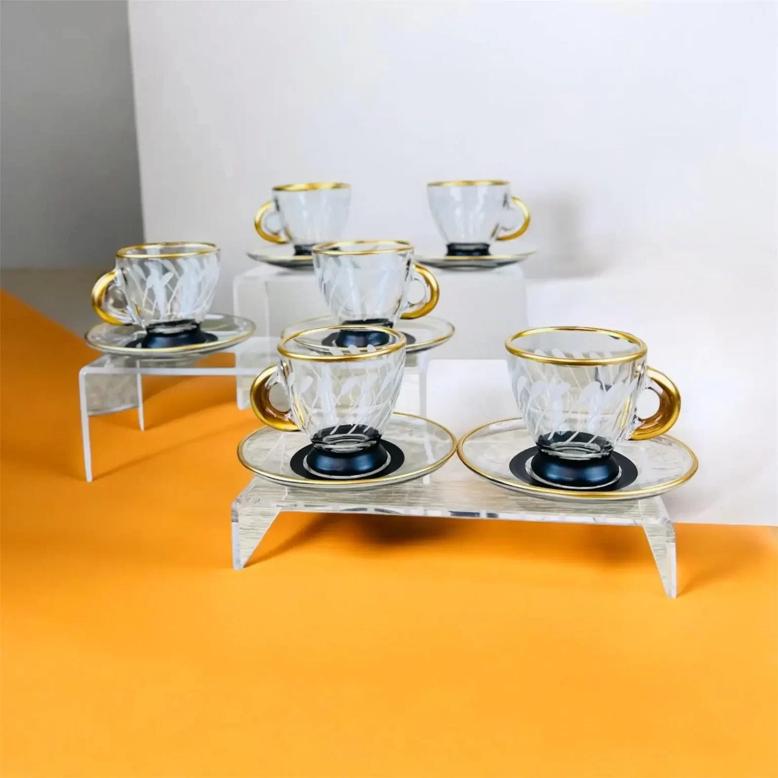 Uppsättning av sex eleganta glaskoppar med guldkant och skålar, visas på ett akrylställ mot en orange och vit bakgrund.