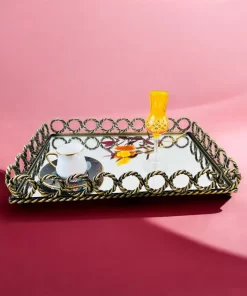 Kvadratisk speilbrett med dekorativ metallkant i flettet design, med en hvit kopp og et gult glass oppå, plassert mot en rosa bakgrunn.