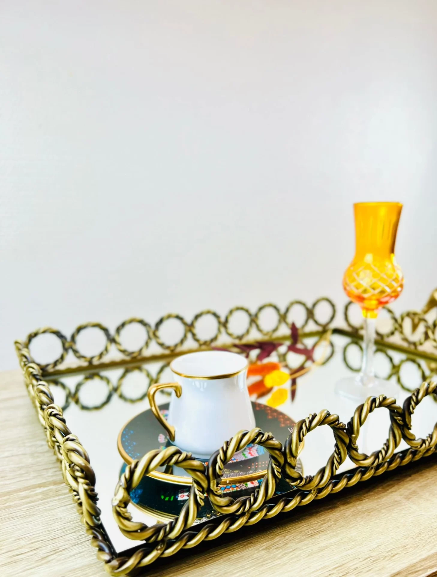 Kvadratisk speilbrett med dekorativ metallkant i flettet design, med en hvit kopp og et gult glass plassert oppå, mot en rosa bakgrunn.