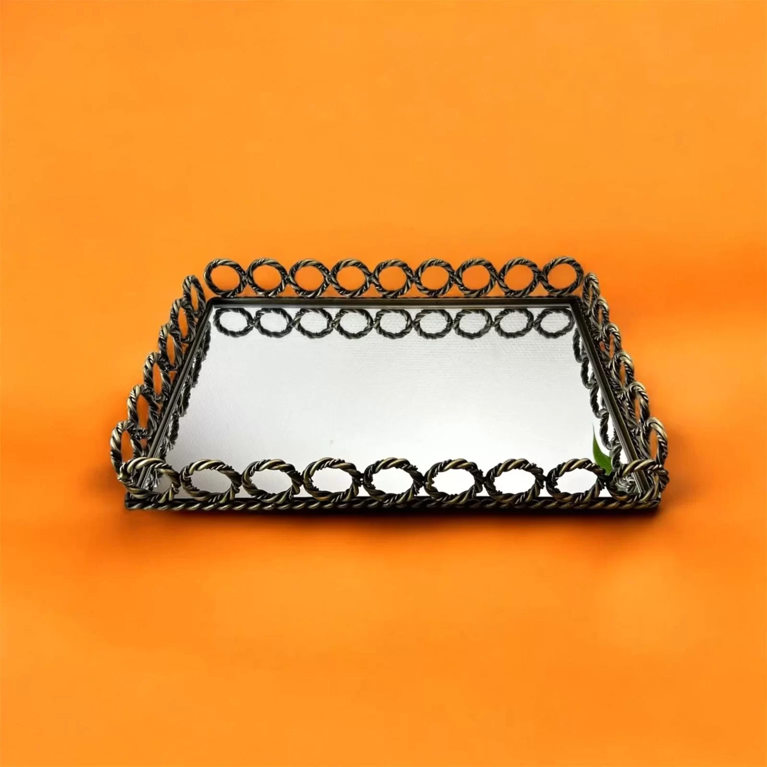 Rektangulär spegelbricka med dekorativ metallbård i flätad design, placerad på orange bakgrund.