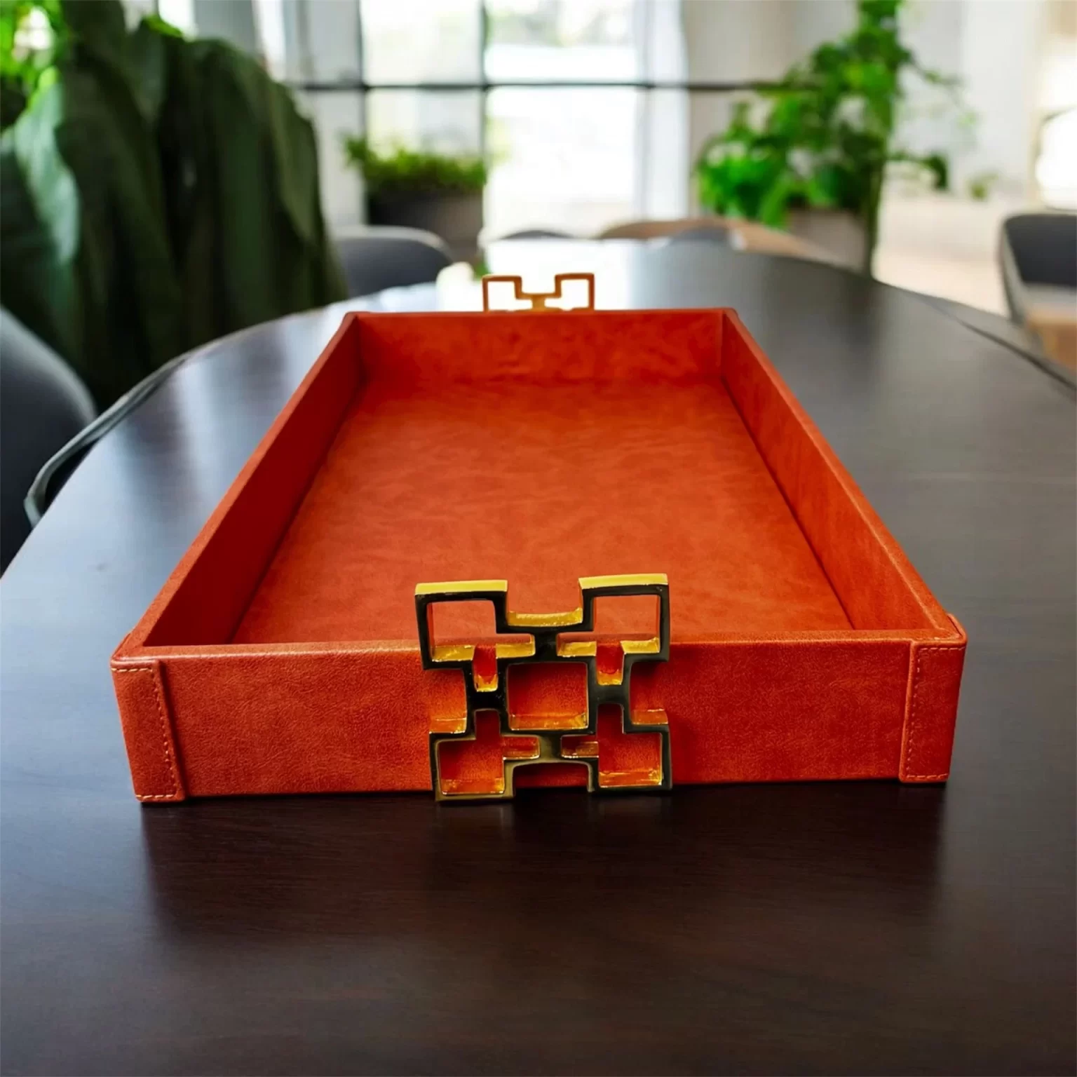 Rektangulær orange serveringsbakke med guldfarvede geometriske håndtag, placeret på et mørkt bord i et lyst rum med grønne planter i baggrunden.