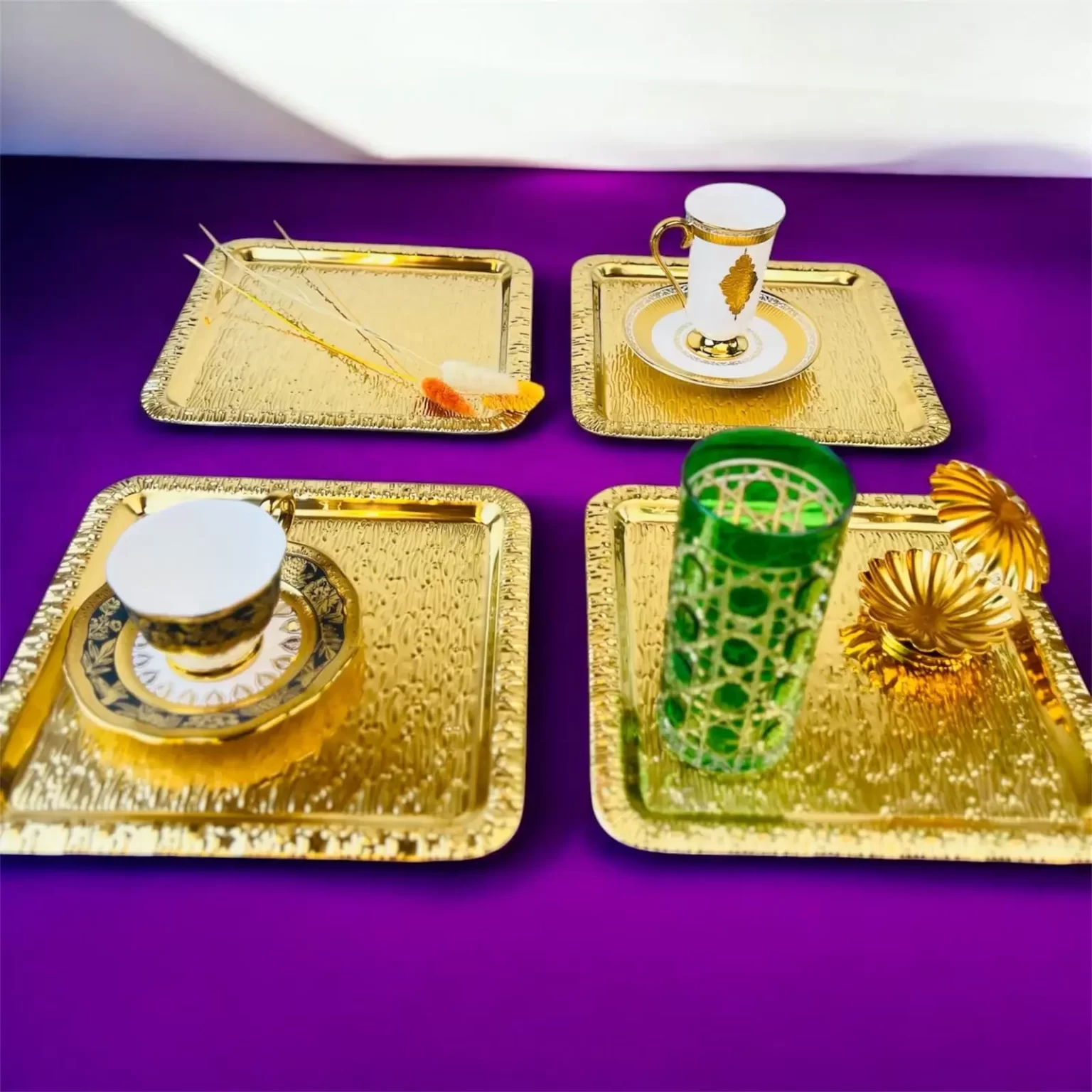 Fire firkantede gullfargede serveringsbrett med kopper, glass og dekor plassert på en lilla overflate.
