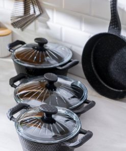 Tre svarte gryter med glasslokk og en stekepanne fra Acar står på en lys kjøkkenbenk foran hvite fliser.